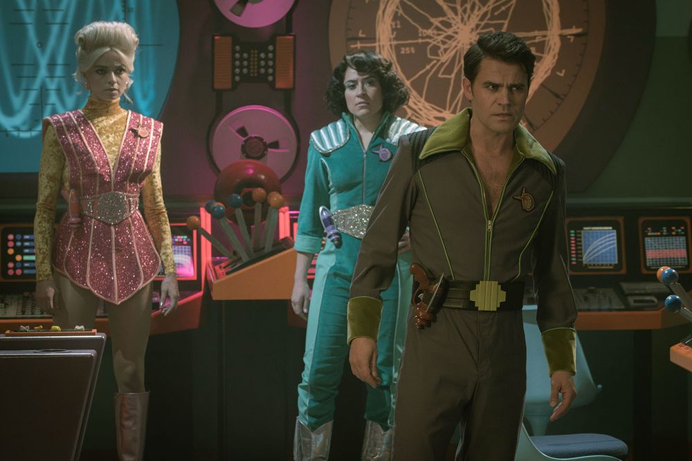 A promo image of Star Trek: Strange New Worlds “A Space Adventure Hour”. On the left is Jess Bush wearing a pink and gold sparkling outfit. In the middle is Melissa Navia seeing a flashy teal uniform, and on the right is Paul Wesley wearing a more subdued green uniform with zipper half zipped and a belt with a laser pistol on his side. 