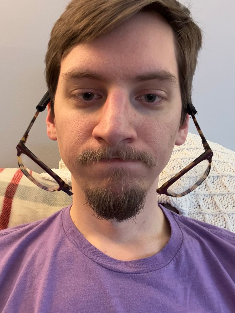 A selfie of Caleb with disappointed expression on his face. His glasses are broken in half and dangling from his ears. 