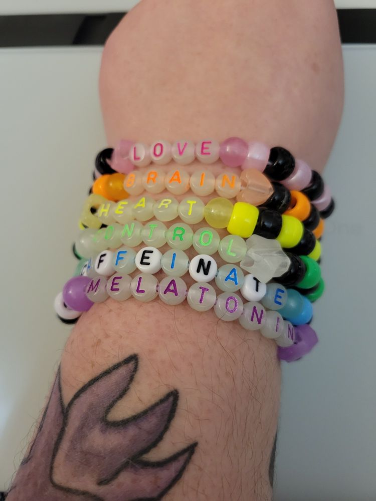 Six bracelets with the separate words "Heart, Brain, Love, Control, Caffeinate, Melatonin" on each one. The style of bracelet is called "kandi", each bracelet has black beads and a colour of the rainbow. Bracelets are on a wrist with tattoos below them