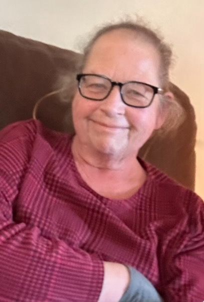 Picture of a older middle-aged woman. Her hair is frizzy and greyish-blonde. She is wearing black glasses and has a smile on her face. Her sweater is a burgundy plaid. The picture is taken at a slight angle, and is a bit out of focus.