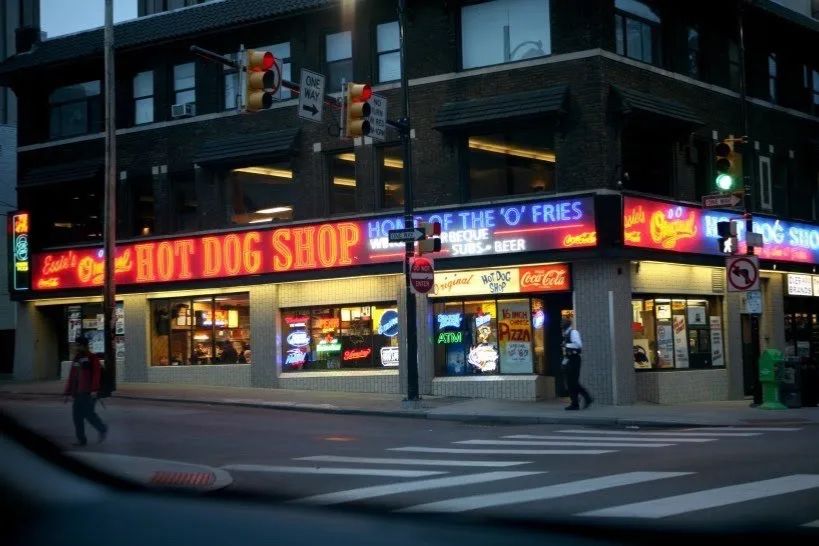 Red neon sign on a city building, street level, reading "the original hot dog shop" next to a blue neon sign reading "home of the "o” fries