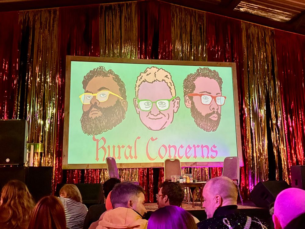 Pre-show set of the stage at Fairfield Social Club. Glittery curtain and a screen with hand drawn faces is Chris Cantrill, Sunil Patel and James Shakeshaft
