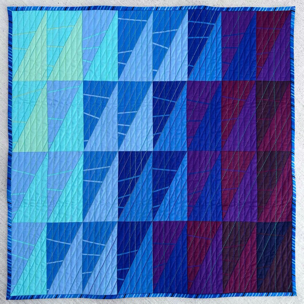 24" x 24"

Description: This mini quilt uses half-rectangle-triangles (HRT) in colors ranging from pale green to deep violet to evoke the colors of the sea. The color moves forward and back in an uneven gradient to emulate the movement of water in a geometric form. Solid fields of color are broken with 1/8" wide pieced slivers in one-half of each HRT unit.