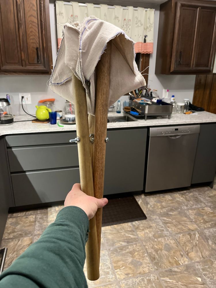 Folded up camping stool with three wooden legs and a white cloth seat. The three legs are held in a single hand.