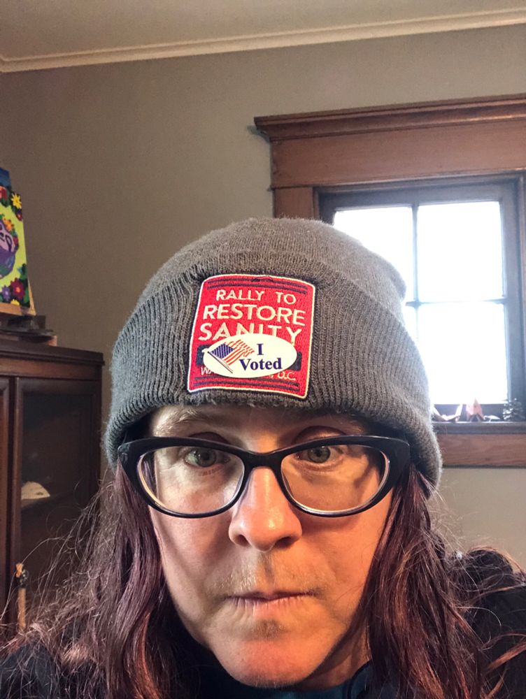 A female figure with long purple hair, close to the camera, wearing glasses and a grey knit beanie. The female figure is not smiling. Her had has an ‘I voted’ sticker stuck to the forehead.