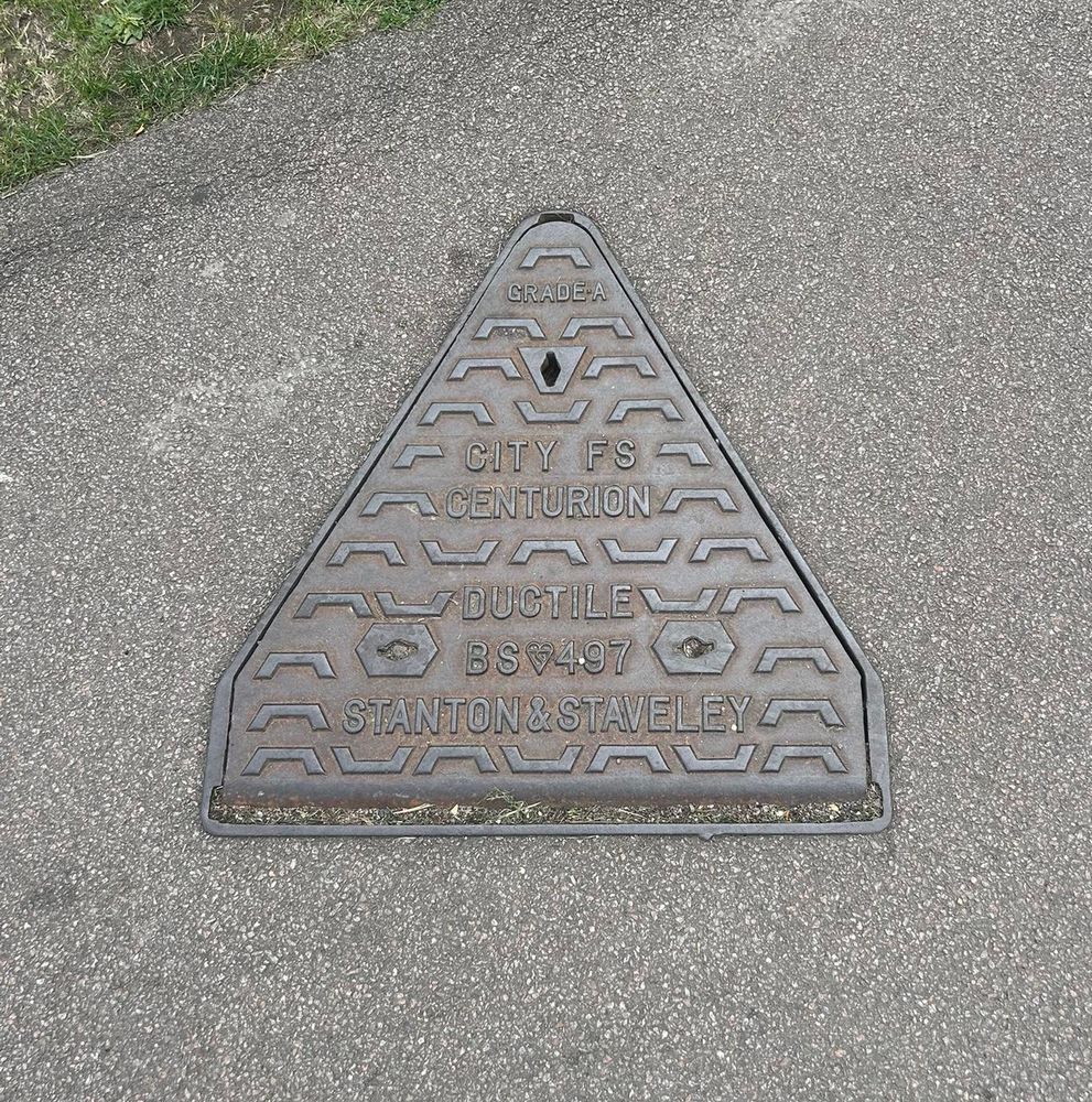 Photo of a triangular-shaped manhole cover in a tarmac footway.
Lettering on it says
Grade A
City FS Centurion
Ductile
BS497
Stanton & Staveley
