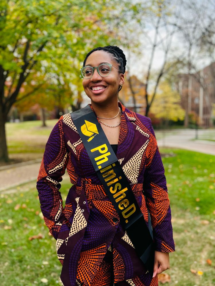 Woman with a printed suit stands smiling wearing a sash that says “Ph.inisheD.
