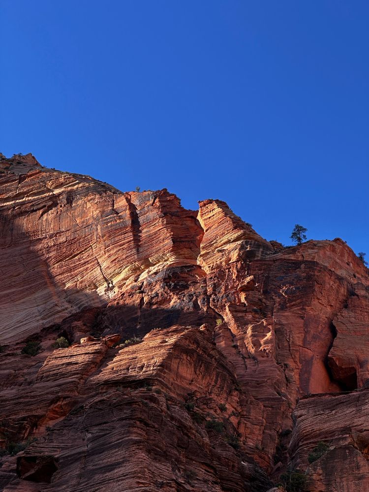 An upward view from the very foot of a red cliff face. The cliff has wavy horizontal grooves etched into the very face of the rock, revealing layers of colours and textures. Above it hangs a clear azure sky.