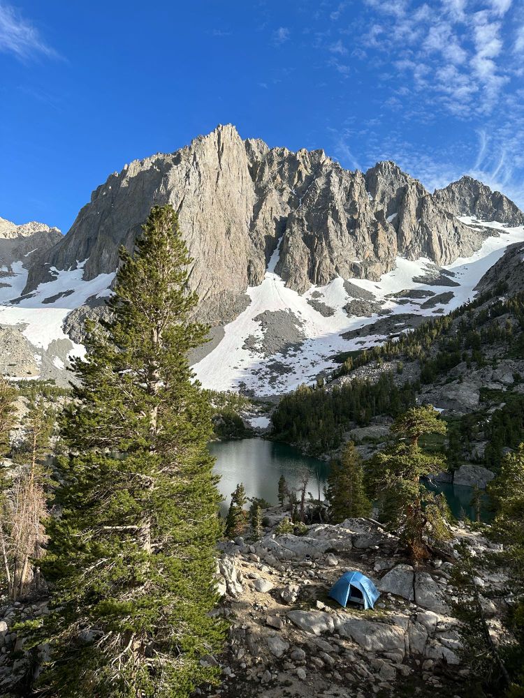 The Palisades in the Sierra Crest: jagged snow-footed peaks lining the background against a bright blue sky. The midground has a dark emerald lake ("Third Lake") at the foot of the peaks. My blue tent and backpacking camp is set up in the foreground- looking diminutive against the soaring peaks behind.