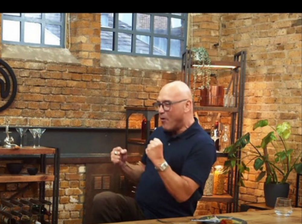 Gregg of master chef standing behind a table in the studio. He wears a blue polo and he holds his arms vaguely boxer pose. His trousers are dark brown and you can see one knee so he is standing on one leg and the other leg bend before him. Fighter pose