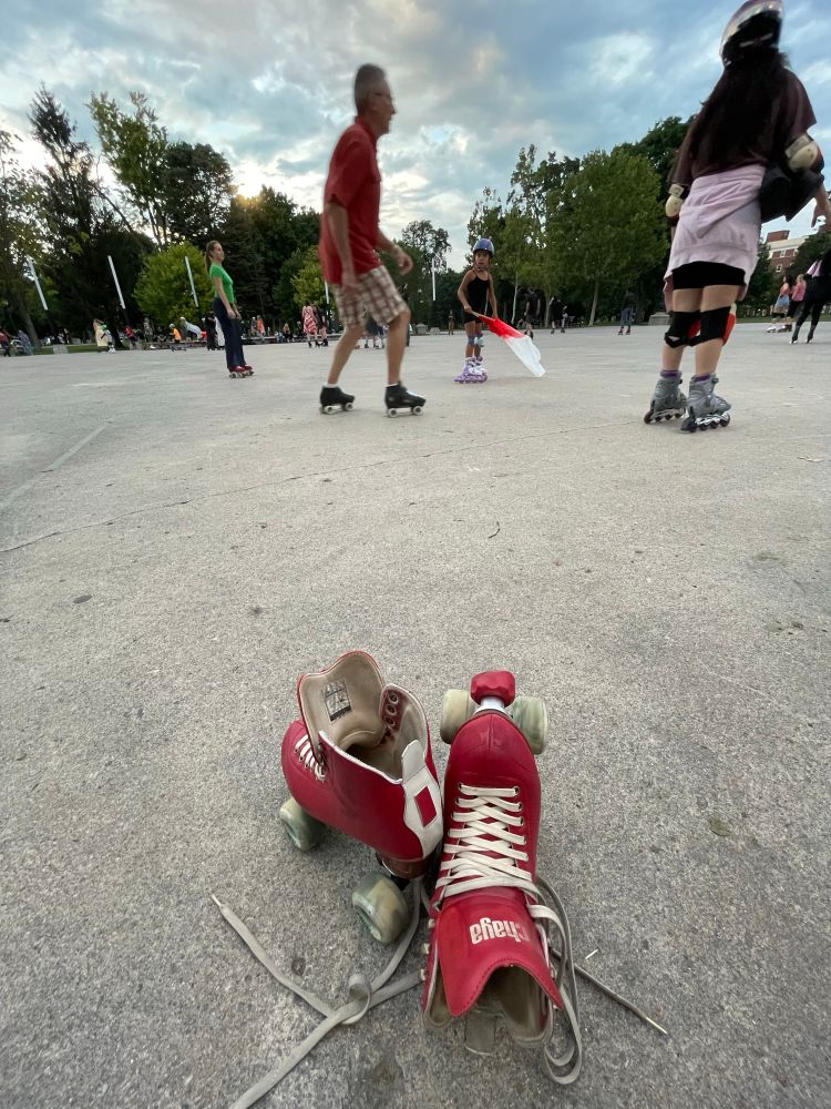 A pair of red roller skates sit on pavement in the foreground. People roller skating. Trees in the background with a partially couldn’t sky. 