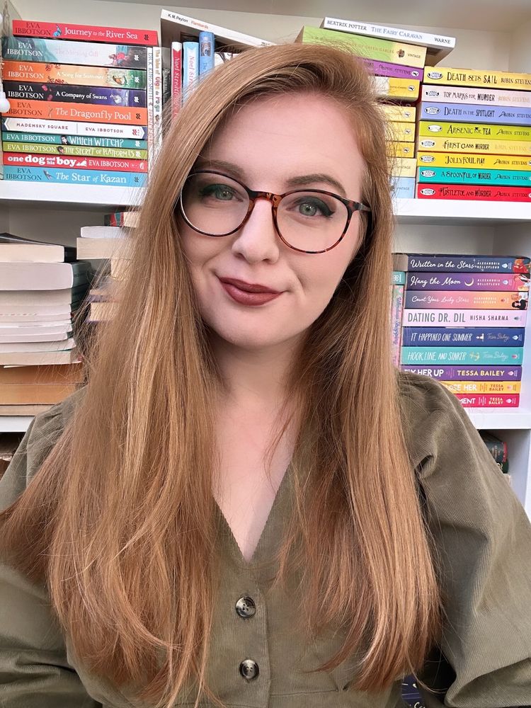 Lucy, a white woman in her twenties, with strawberry blonde hair, round glasses, winged eyeliner, lipstick, and wearing a green dress. Taken in front of a full, colourful bookshelf with stacked books. 