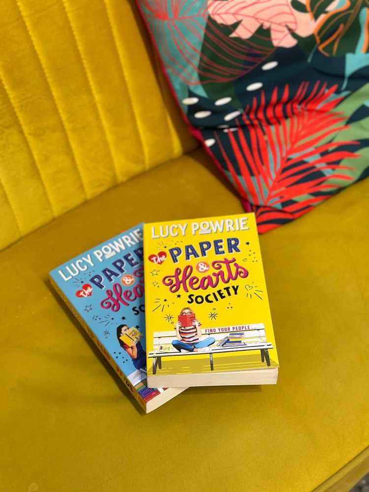 The Paper & Hearts Society and Read with Pride by Lucy Powrie, books sitting on a yellow sofa. 