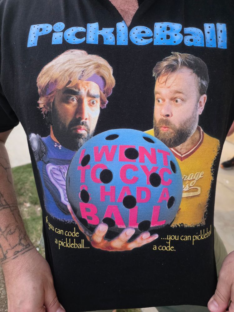 Close-up of a novelty black T-shirt with the word 'PickleBall' at the top in blue letters. The shirt features two illustrated faces of men looking surprised at a large blue pickleball in the center. The ball has pink text that reads, 'I WENT TO CYC & HAD A BALL.' Below, the tagline says, 'If you can code a pickleball… you can pickleball a code.'