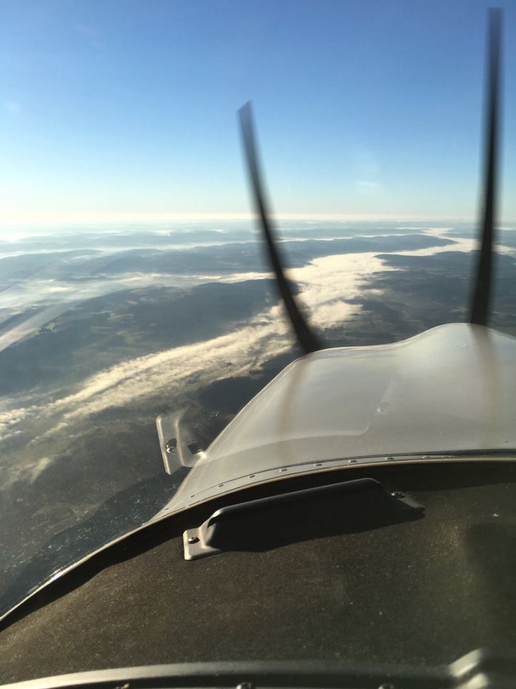 This photo appears to be taken from the cockpit of a small airplane, looking out over a scenic landscape. The spinning propeller blades are visible in the foreground, creating a blurred effect due to their motion. Below, the landscape shows rolling hills or mountains partially covered by a layer of mist or fog, under a clear blue sky. The perspective highlights the freedom and vastness often experienced during flight.