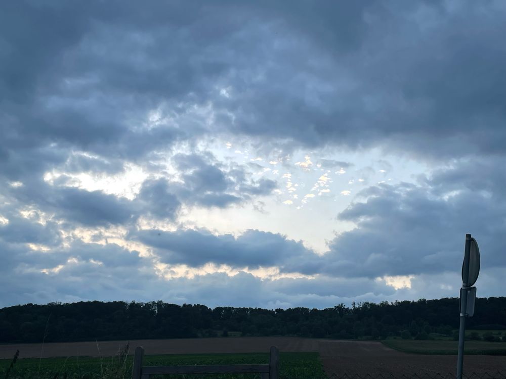 Foto eines abend-Himmels, in der Mitte ist ein Loch in der Wolkendecke, hinter dem man noch Sonnenschein und einzelne kleine, höhere Wölkchen sieht. Am unteren Bildrand sieht man einen schon dunklen Waldrand u d darunter einige Felder.