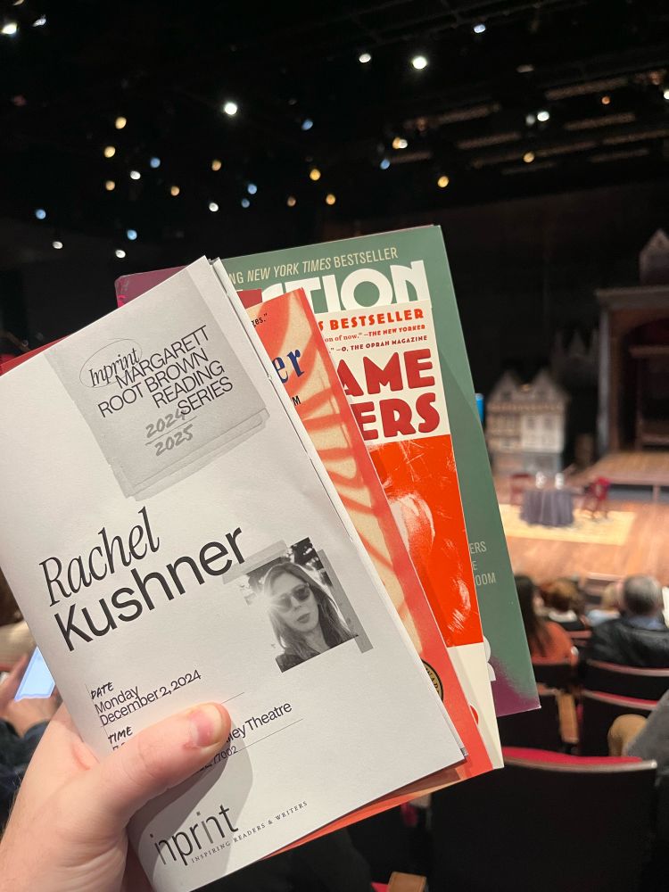 A program for a reading with Rachel Kushner along with 3 of her books. The books are being held up by someone in a theater seat with the theater positioned out of focus in the background. 