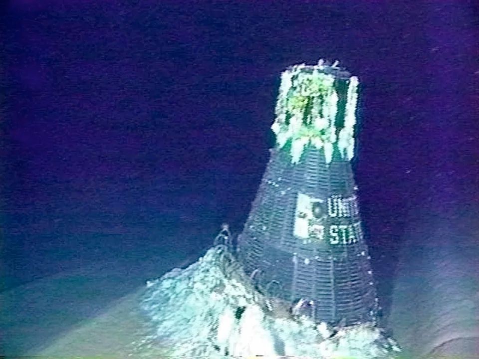 Another more distant view of the other side of Mercury spacecraft, with painted with "United States" in white, photographed on the abyssal ocean plain. The craft is encrusted with corals and other marine life and is partially embedded in soft sediments.