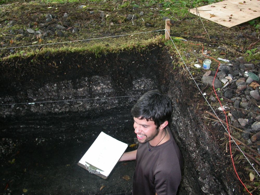 Jacob a handsome young archaeologist  in a two metre deep archaeological excavation which shows the mossy forest floor underlain by thick deposits of shell midden, which shows as speckled white and gray dirt with horizontal layers. pic by qmackie