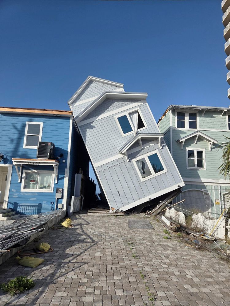 A photograph of three houses in a row, the middle one has fallen off stilts and sits at a sharp angle leaning on both houses to the sides.