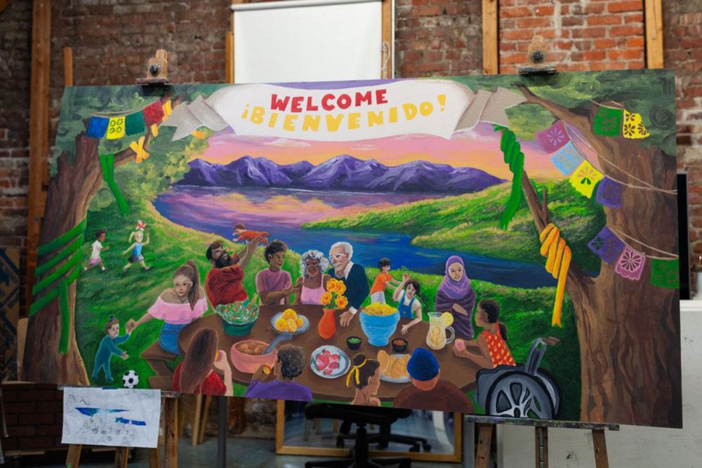 A painted mural depicts a diverse group of people of all ages sitting around a table of food outdoors with a range of purple mountains and pink sky reflecting in water in the background. Colorful flags hang in the trees. A banner reads: "Welcome / Bienvenido" 
