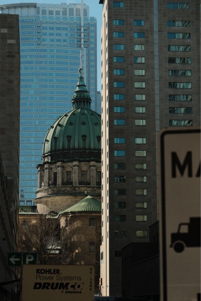 The photograph shows an old downtown Montreal church trapped between tall 20/21st century towers. The picture is zoomed, so you can see in the foreground part of a sign for trucks, and a packing sign in the middle ground. It’s like a layer of building and the church seems very small compared to the rest.