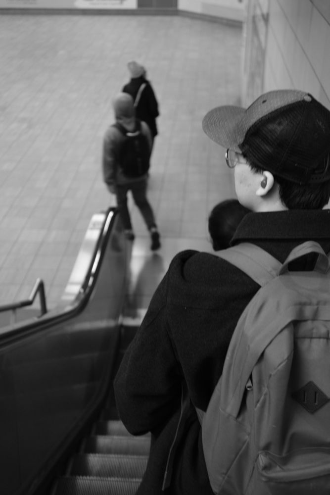 A person wearing a backpack is descending into a train station on an escalator while looking at people walking below.