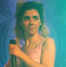 a singer (Marina) holding a microphone stares off in the distance with a slight smile and wide, surprised eyes
