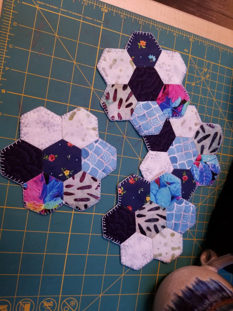 Four coasters made of 6 hexagons around a central hexagon, all sitting on top of a green and yellow cutting mat. one is laying on it's own, and the other three are overlapped. They are made jp of white, black, navy blue, colorful, and printed fabric. They are edged in a light blue.