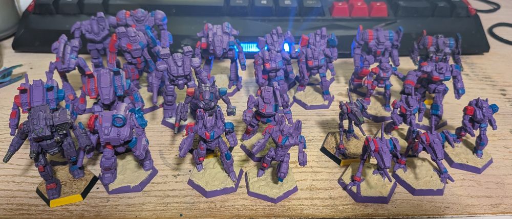 An assortment of Battletech miniatures painted in the colors of the Marik Militia (Purple, blue, and red). Most of the miniatures are half painted. 