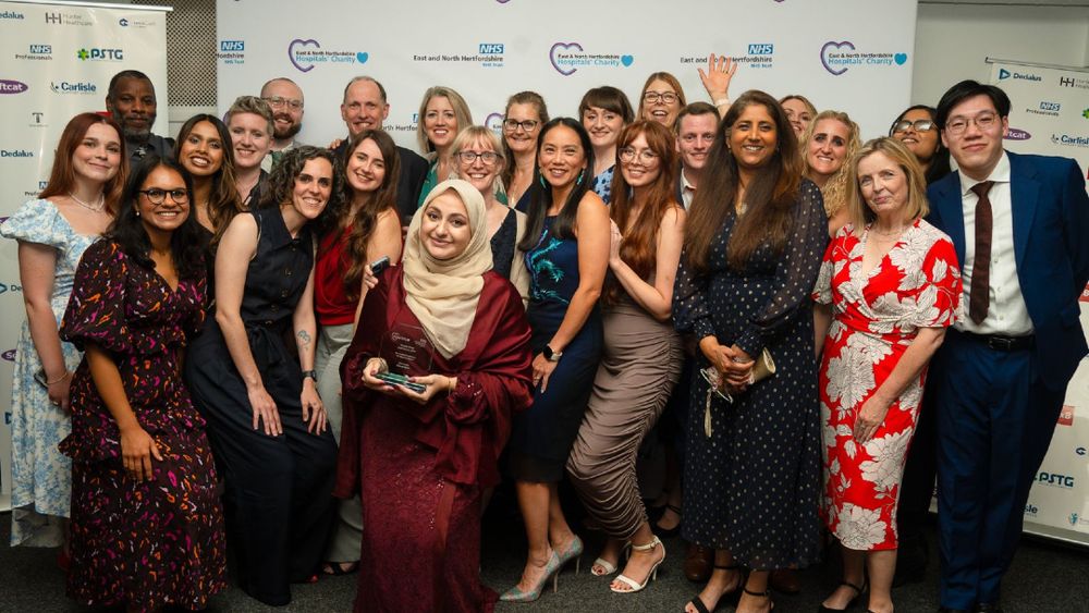 A team of people stand in front of a banner with Trust and charity logos, at the 2024 Time to Shine Staff Awards