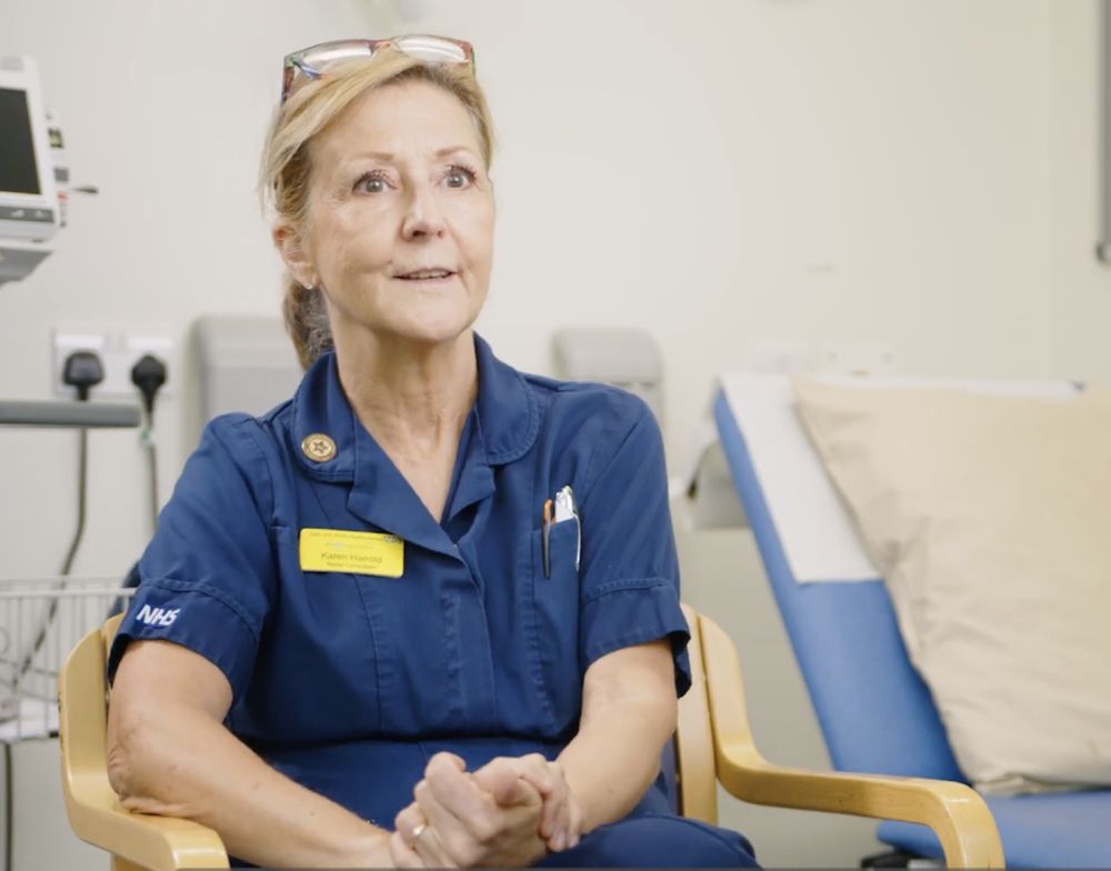 A photograph of Dr Karen taken as a screenshot from a recent video filmed by East and North Hertfordshire NHS Trust. She is photographed wearing a blue nurses uniform while speaking to camera.