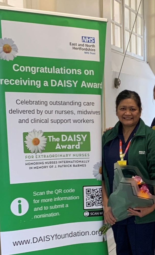 A photo of Cecille holding a bunch of flowers that she received upon winning. Cecille is smiling and standing next to a pull-up banner advertising the DAISY Awards.
