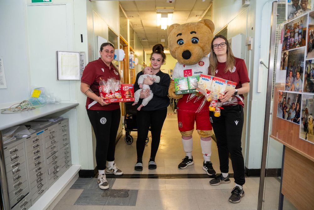 A mother is photographed cradling her young baby on a children's ward at Lister Hospital. She is surrounded by female players from Stevenage Football Club - and club mascot Boro Bear - who are holding chocolate Easter eggs.