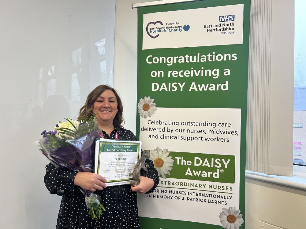 A photo of Penny holding her DAISY Award certificate and a bunch of flowers that she received upon winning. Penny is smiling and standing next to a pull-up banner advertising the DAISY Awards.