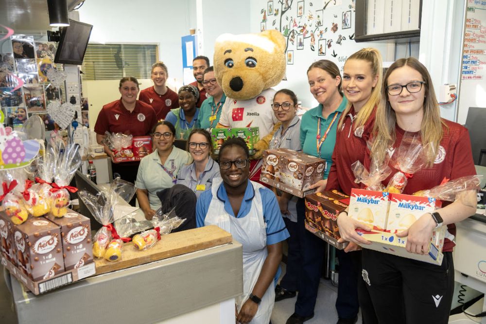 Members of staff on Bluebell Ward at Lister Hospital are photographed alongside players from Stevenage Football Club, as well as the club mascot Boro Bear. They are smiling toward the camera.