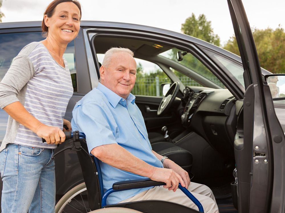An image of a lady collecting an elderly relative in a wheelchair and supporting him into a car. They are both smiling toward the camera.