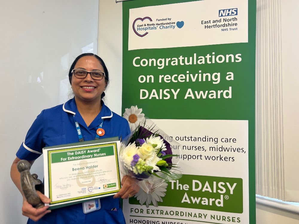 A photo of Beena holding her DAISY Award certificate and a bunch of flowers that she received upon winning. Beena is smiling and standing next to a pull-up banner advertising the DAISY Awards.