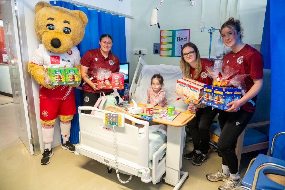 A young patient is photographed in her bed on a children's ward at Lister Hospital. She is surrounded by footballers of the Stevenage Football Club's women's team who are handing her chocolate Easter eggs.