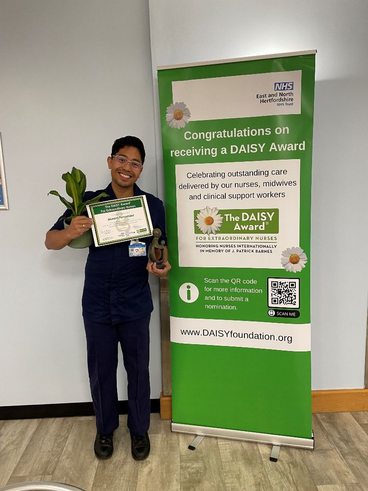 A photo of Akeem holding his DAISY Award certificate and a bunch of flowers that he received upon winning. Akeem is smiling and standing next to a pull-up banner advertising the DAISY Awards.