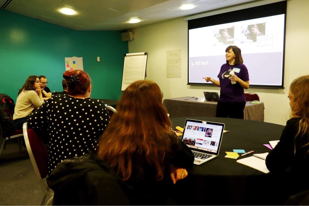 Deborah in a purple MOT T-shirt in front of a projection screen and a small room of people