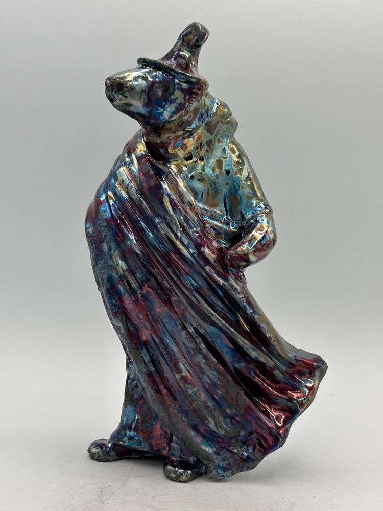 A raku bear sculpture in a witches hat and robe that has been raku fired.  The glaze is iridescent and multicoloured. 