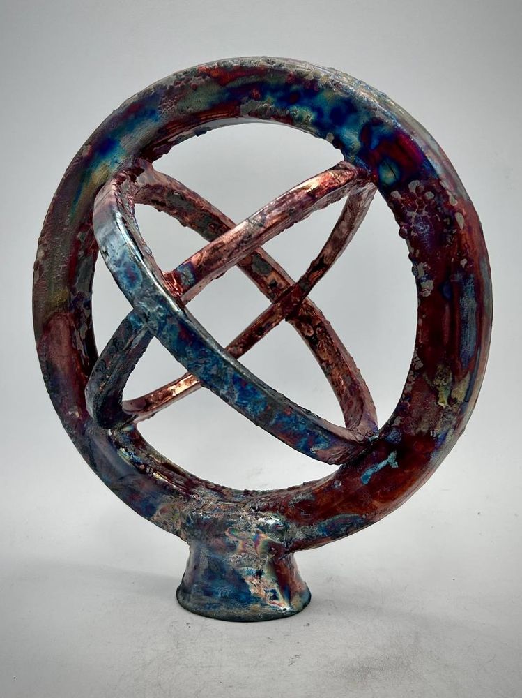 A circular sculpture with two rings inside it. Each ring is to the side of the centre diagonally.  The main circle is on a small base. The colouring is blue, red, copper with a slight rainbow iridescent finish. 