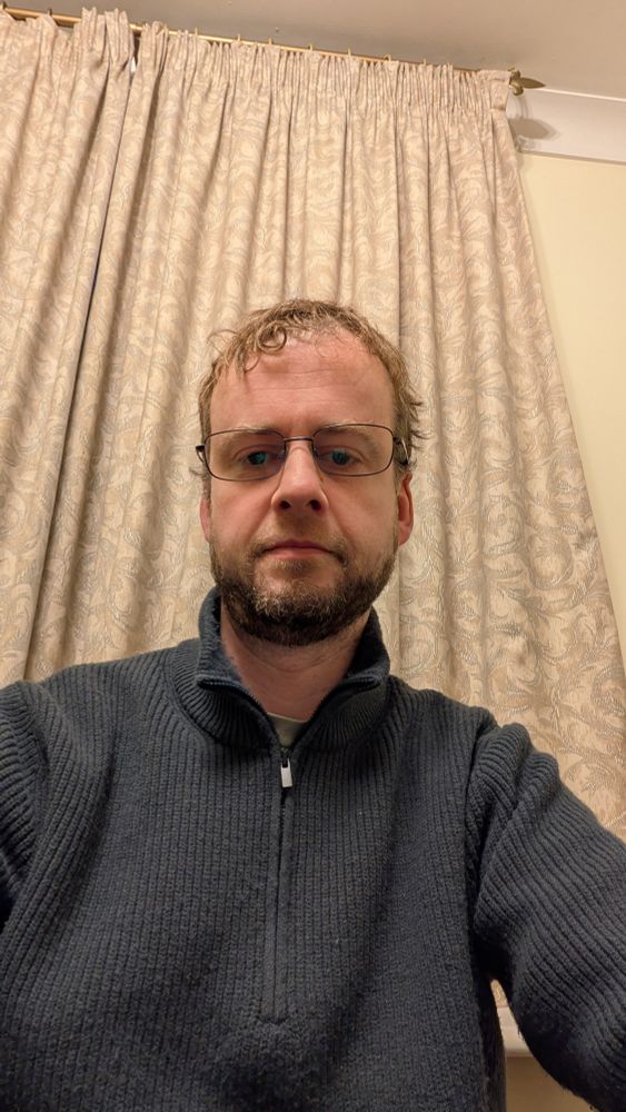 I look into the camera lens wearing a blue jumper with a cream curtain behind me. I am wearing glasses. 