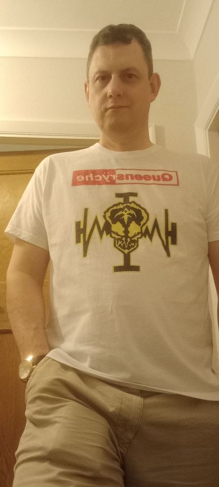 A man wearing a white t-shirt and clay coloured trousers. The t-shirt has the band name Queensryche on it, and a skull with headphones.