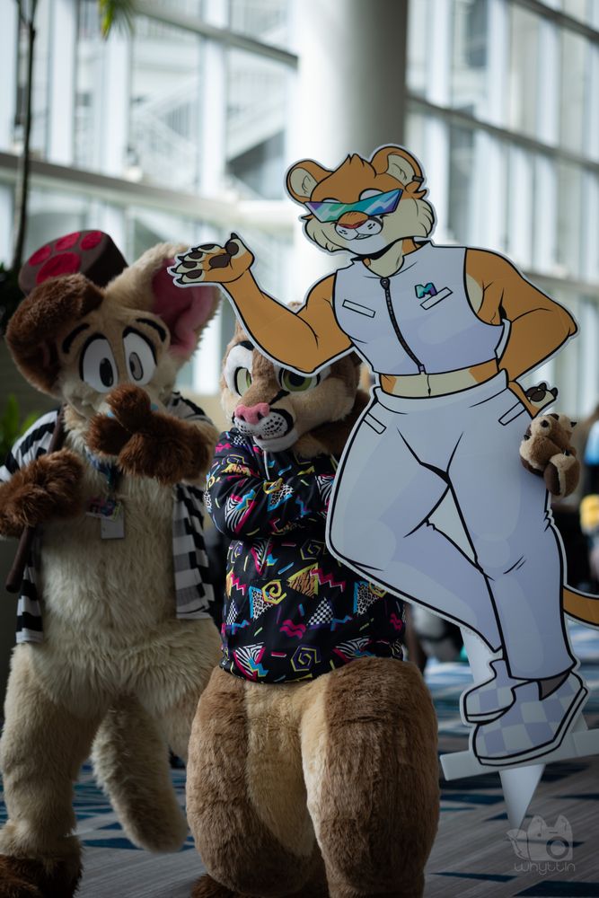 Two full fursuiters walking towards the camera, the tan left one pointing at the camera, the brown and tan right one holding a cutout that was along the parade route.