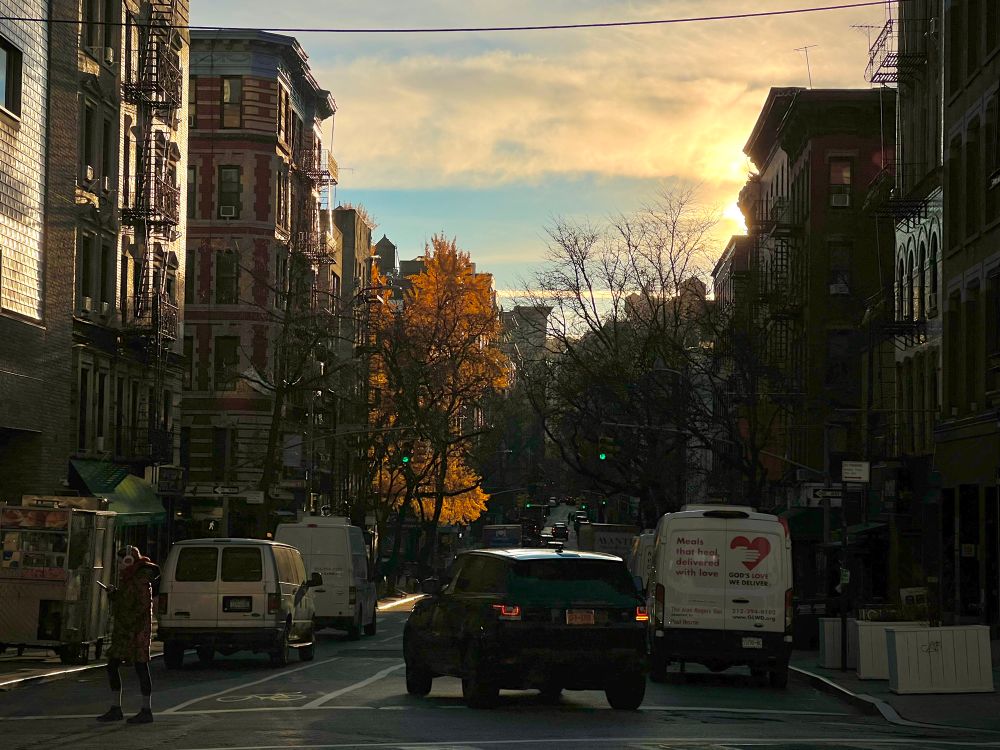 A photo of NY SoHo at 8am. The sun peaks over the rooftops in the right of the frame, casting golden light onto a yellow tree. A car turning centers the frame with a pedestrian crossing to the left.