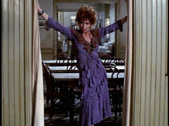 Miss Hannigan from the movie Annie (1982) standing in a doorway with her arms out in a purple dress. 