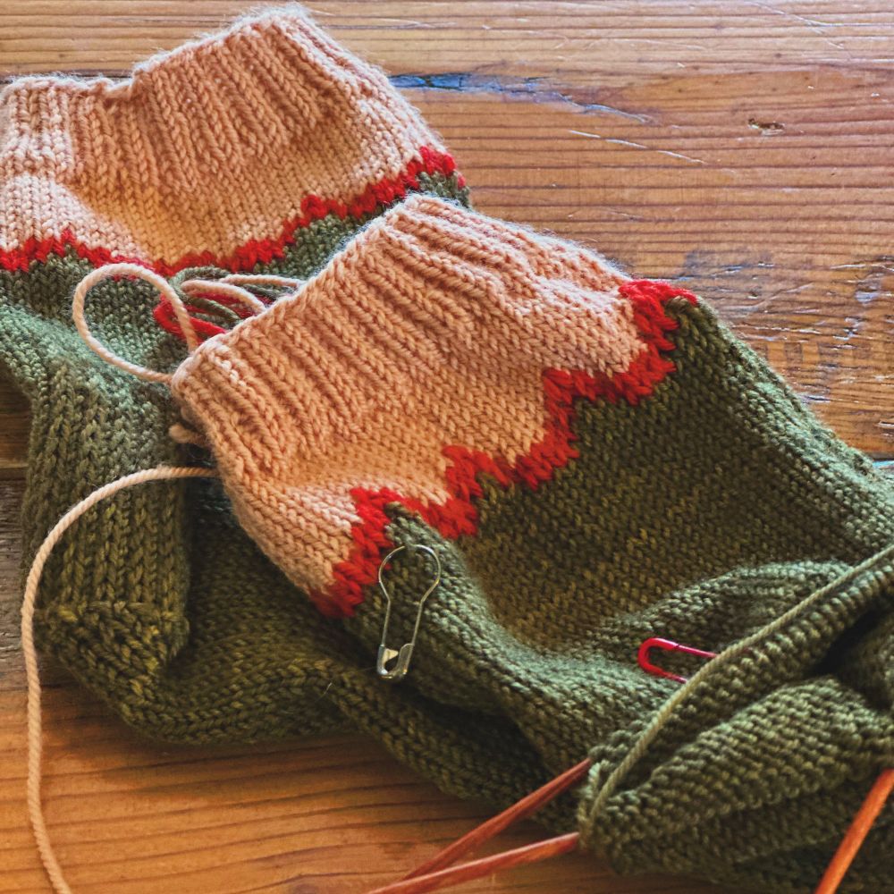 One peach, red, and green sock, plus another half-finished matching sock still on the knitting needles, lying on a wooden table.
