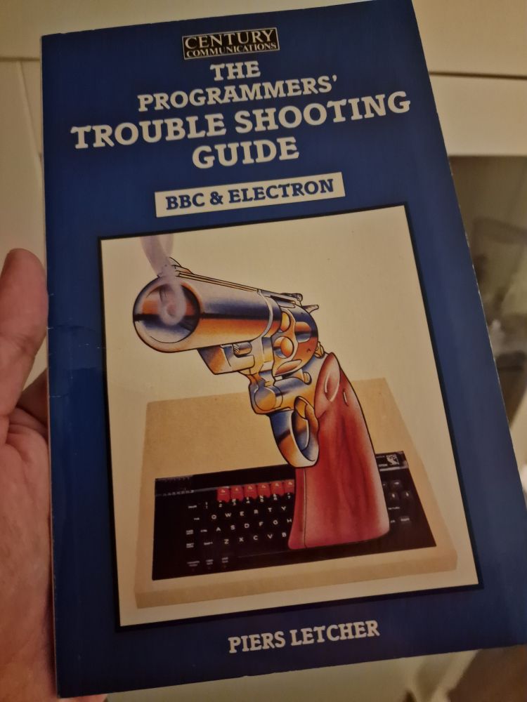Photo of me holding a book called The Programmers' Troubleshooting Guide for the BBC & Electron. Cover depicts a BBC Micro with a huge revolver pointing out towards the reader, barrel smoking.